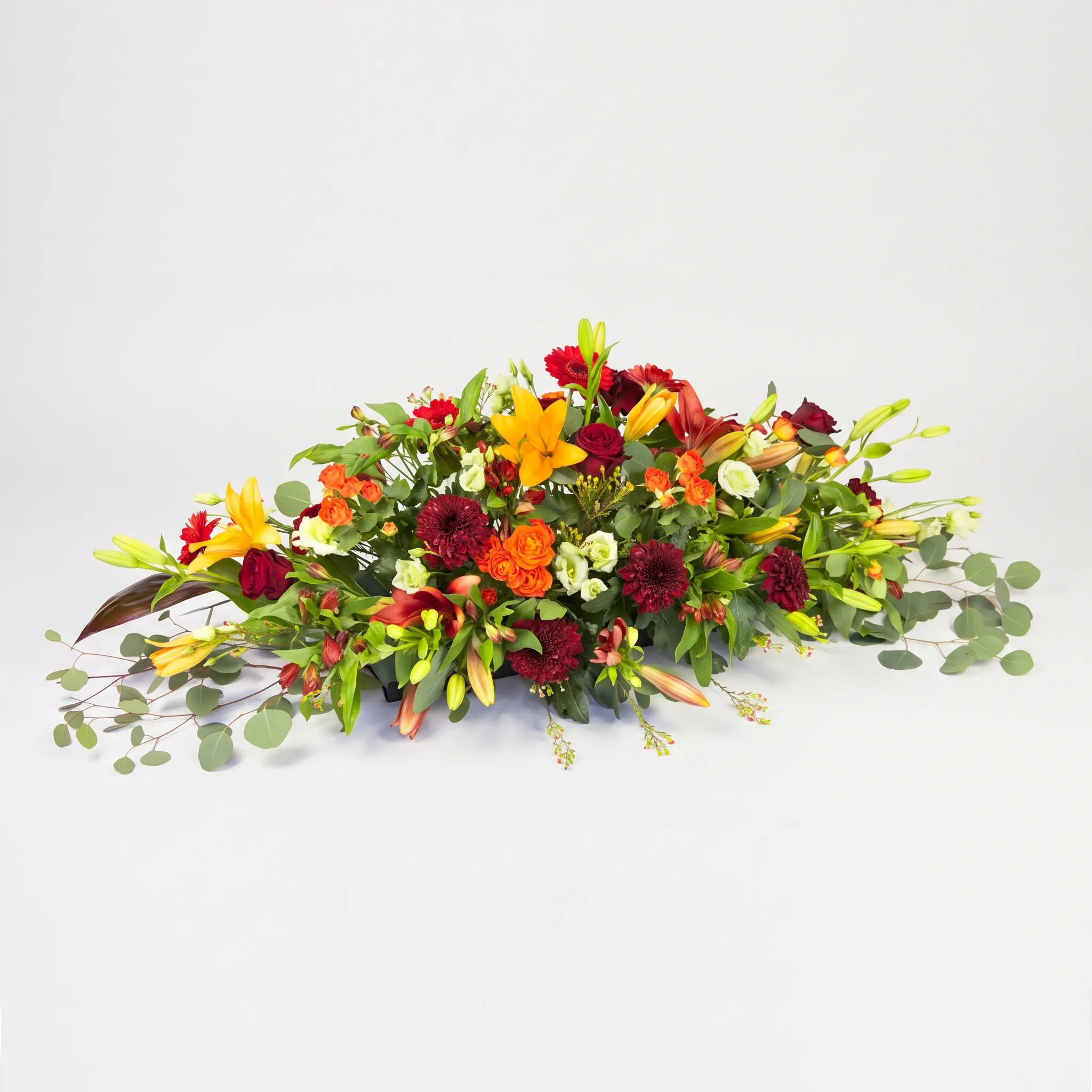 Raquette de cercueil aux tons chauds avec lys jaunes, roses rouges et chrysanthèmes bordeaux